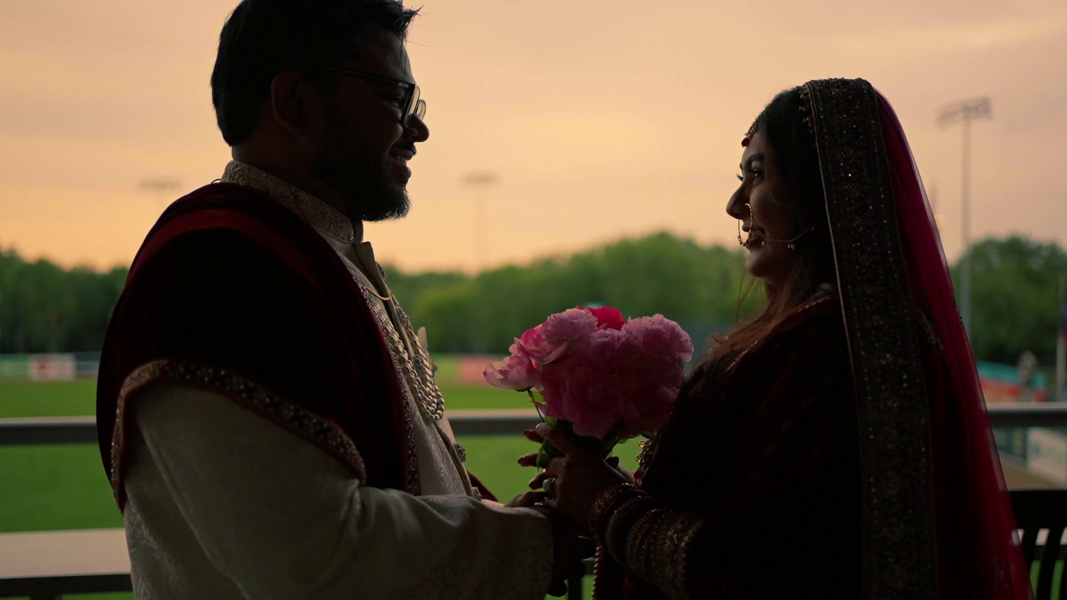 Silhouetted figures share an intimate moment at sunset while holding pink flowers on a porch.