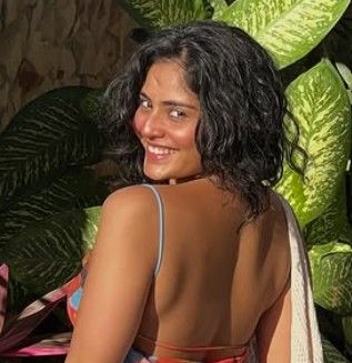 Someone with curly dark hair smiles while looking over their shoulder, with tropical plants in the background.