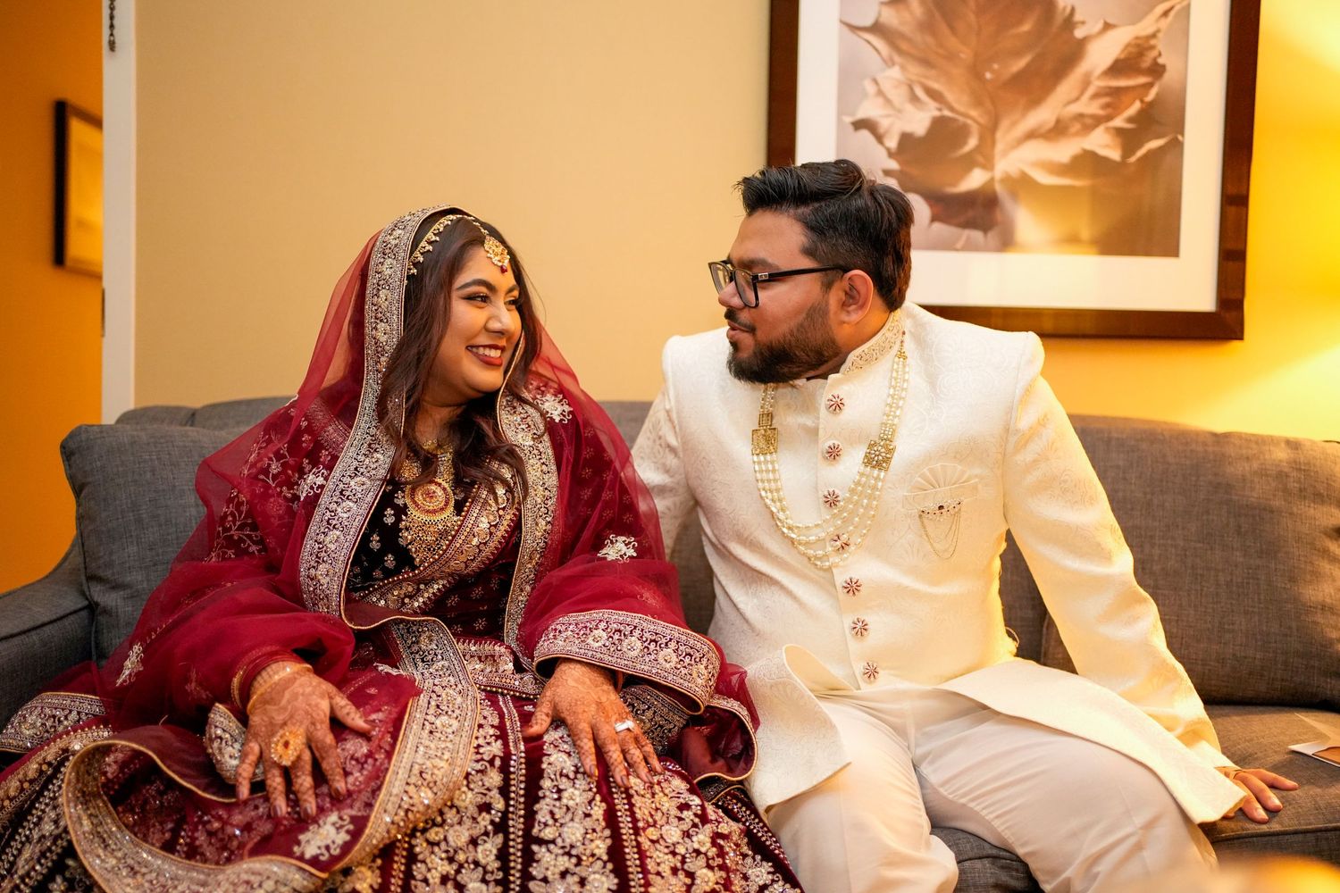 Couple in traditional South Asian wedding attire share a joyful moment while sitting on a grey couch.