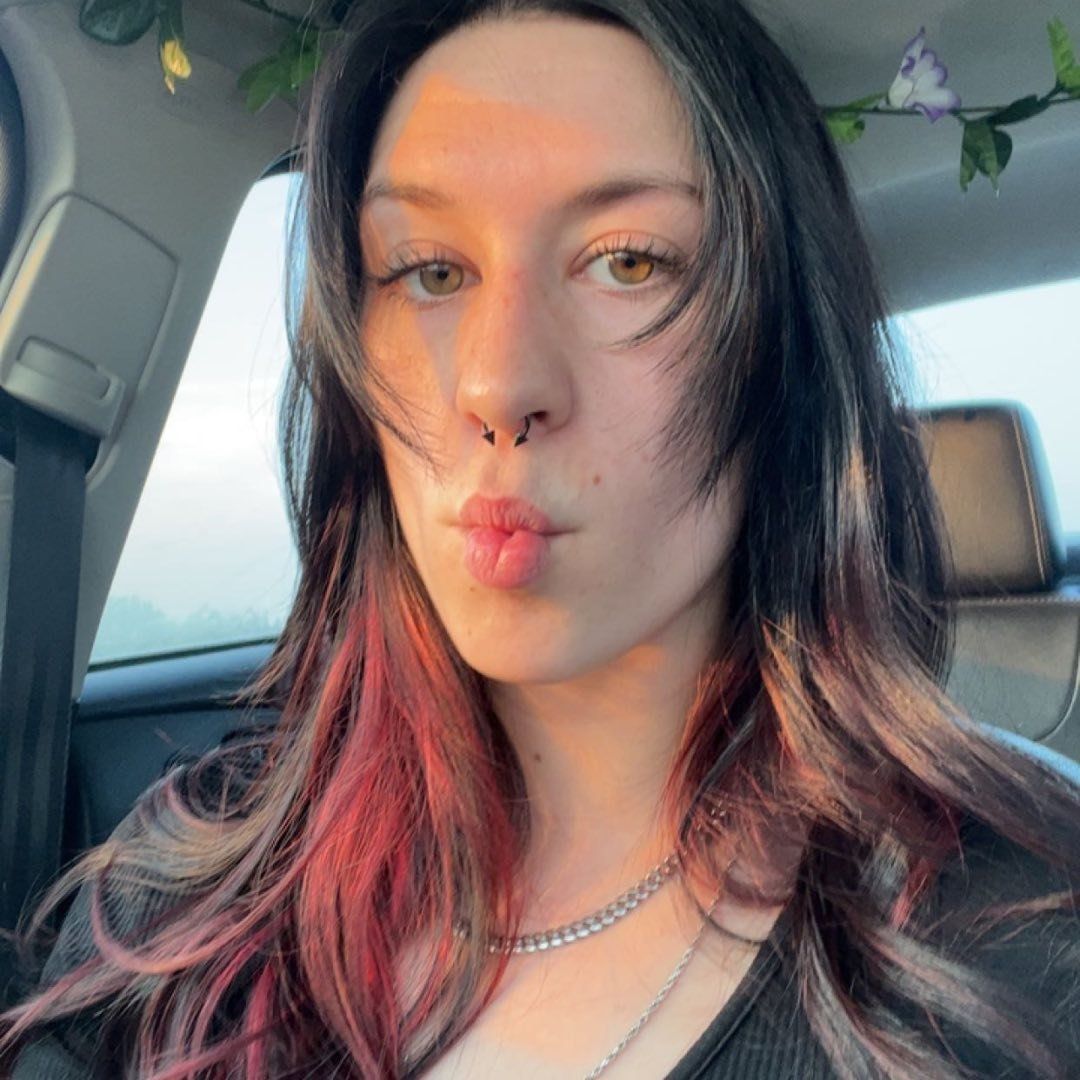A selfie in a car showing long black and red ombre hair, nose piercing, and puckered lips making a kiss expression.