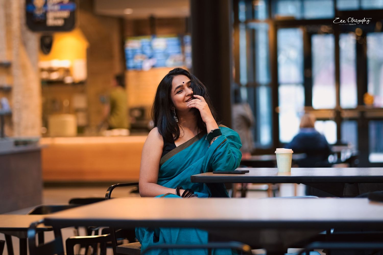 Person in teal saree sitting thoughtfully at a cafe table.