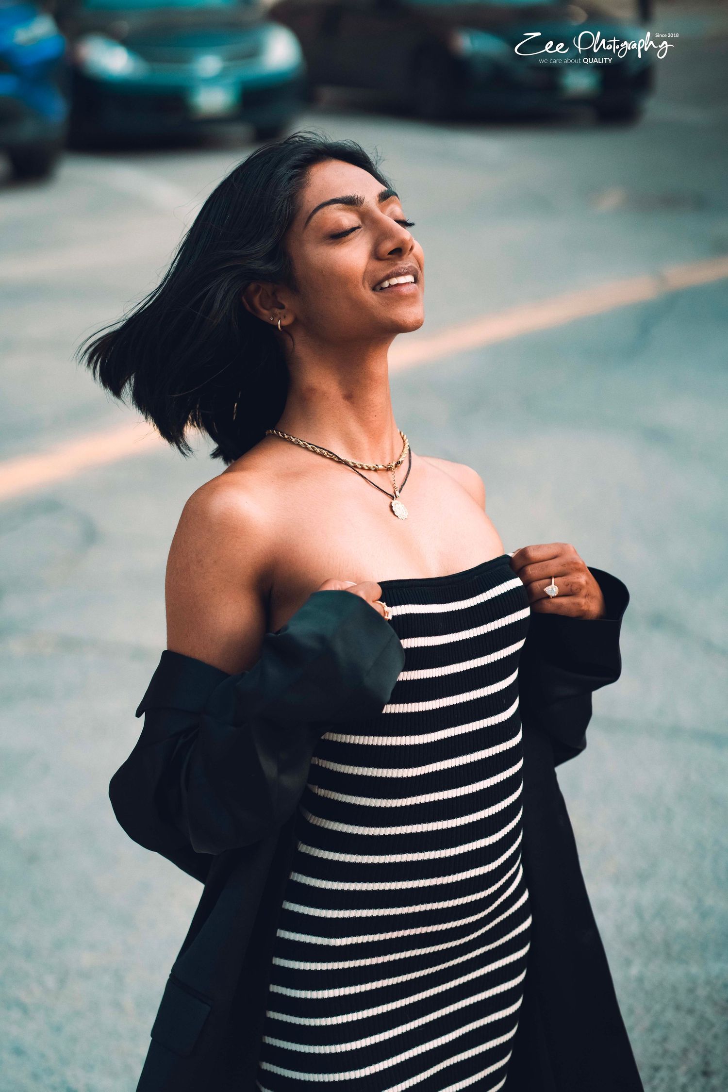 A person in a striped dress and black jacket smiling with eyes closed outdoors.