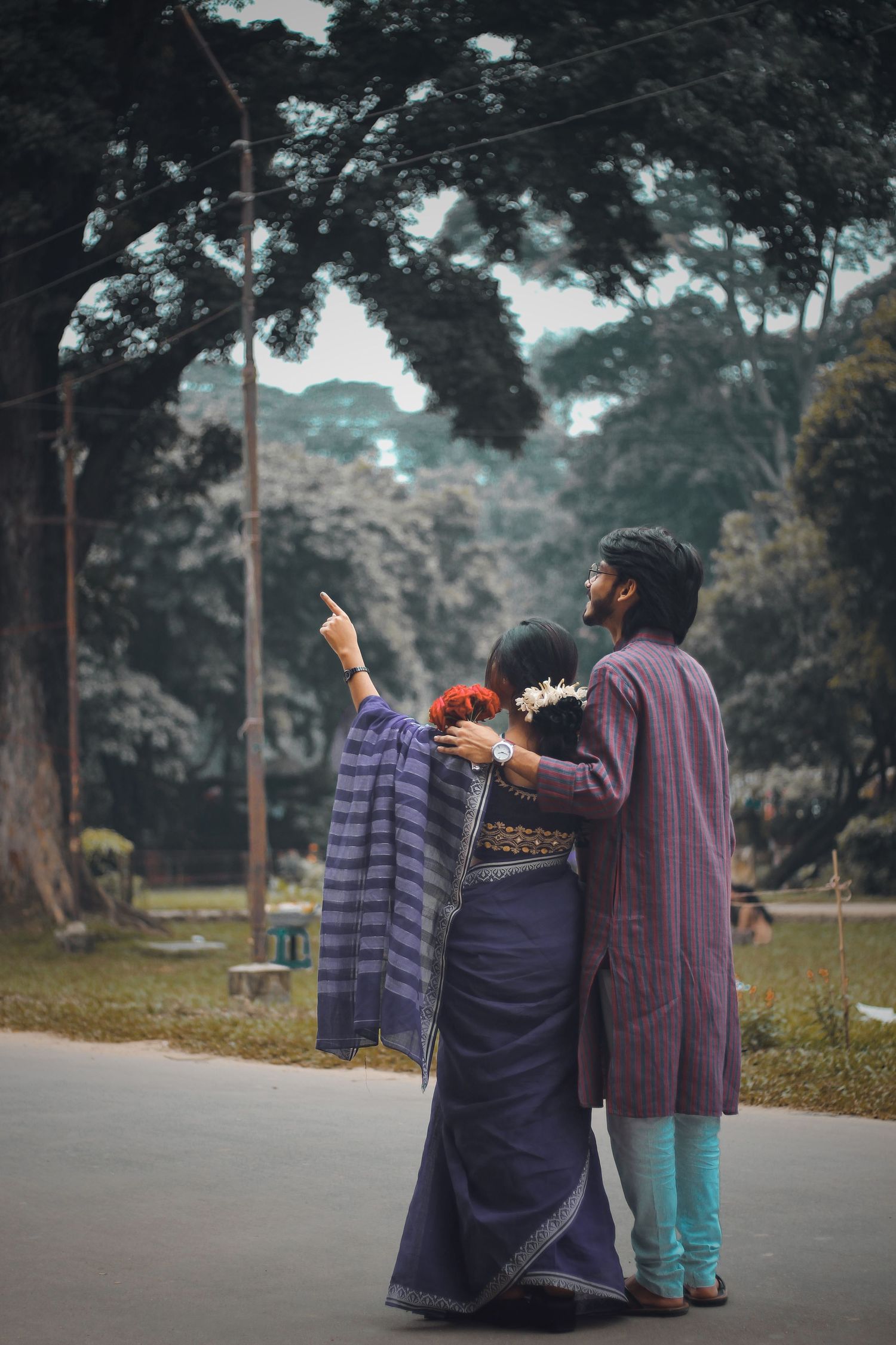 Two people in traditional purple attire standing on a path pointing at something in the distance under tree branches.