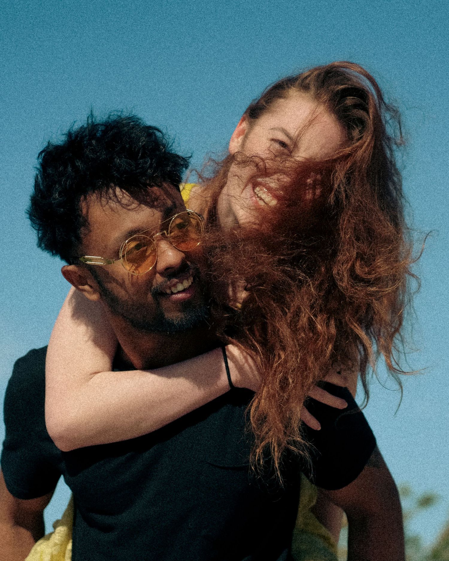 A joyful couple sharing a playful piggyback moment against a blue sky background, both radiating genuine happiness.