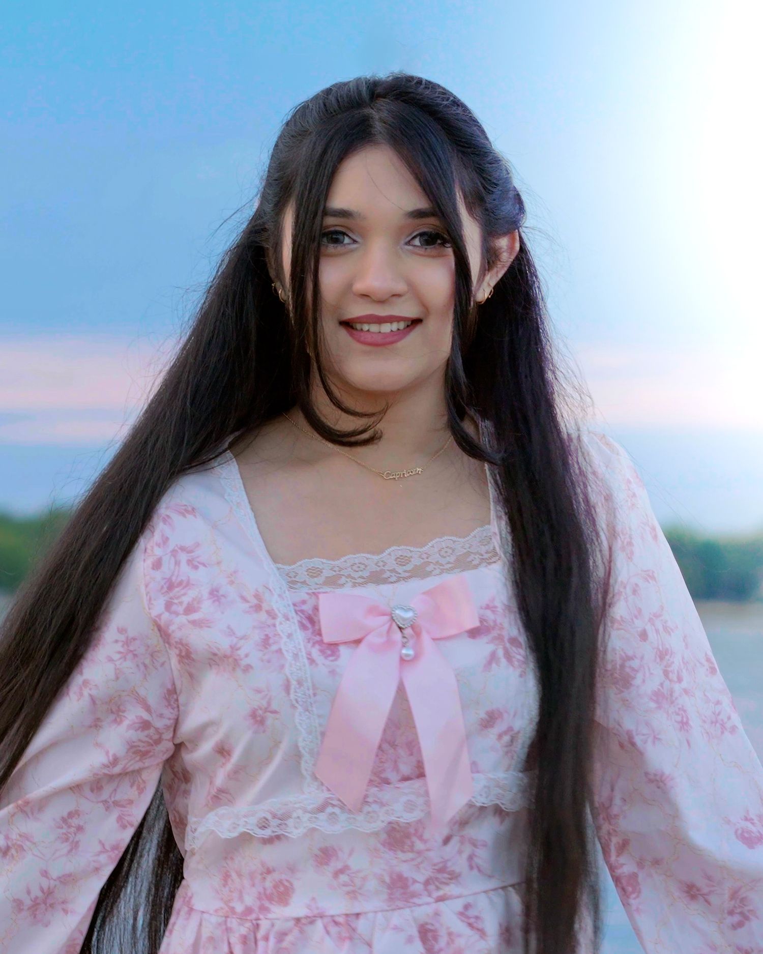 A person with long dark hair wearing a pink floral dress with a bow accent poses outdoors at sunset.