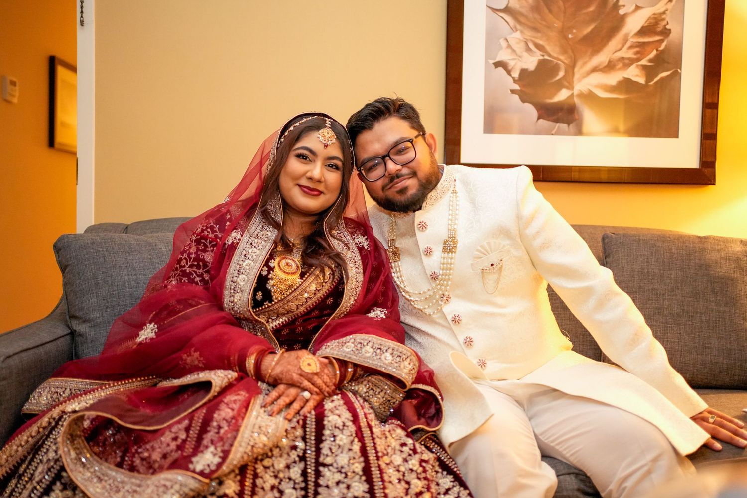 A couple in traditional Indian wedding attire sits together on a gray couch in a warmly lit room.
