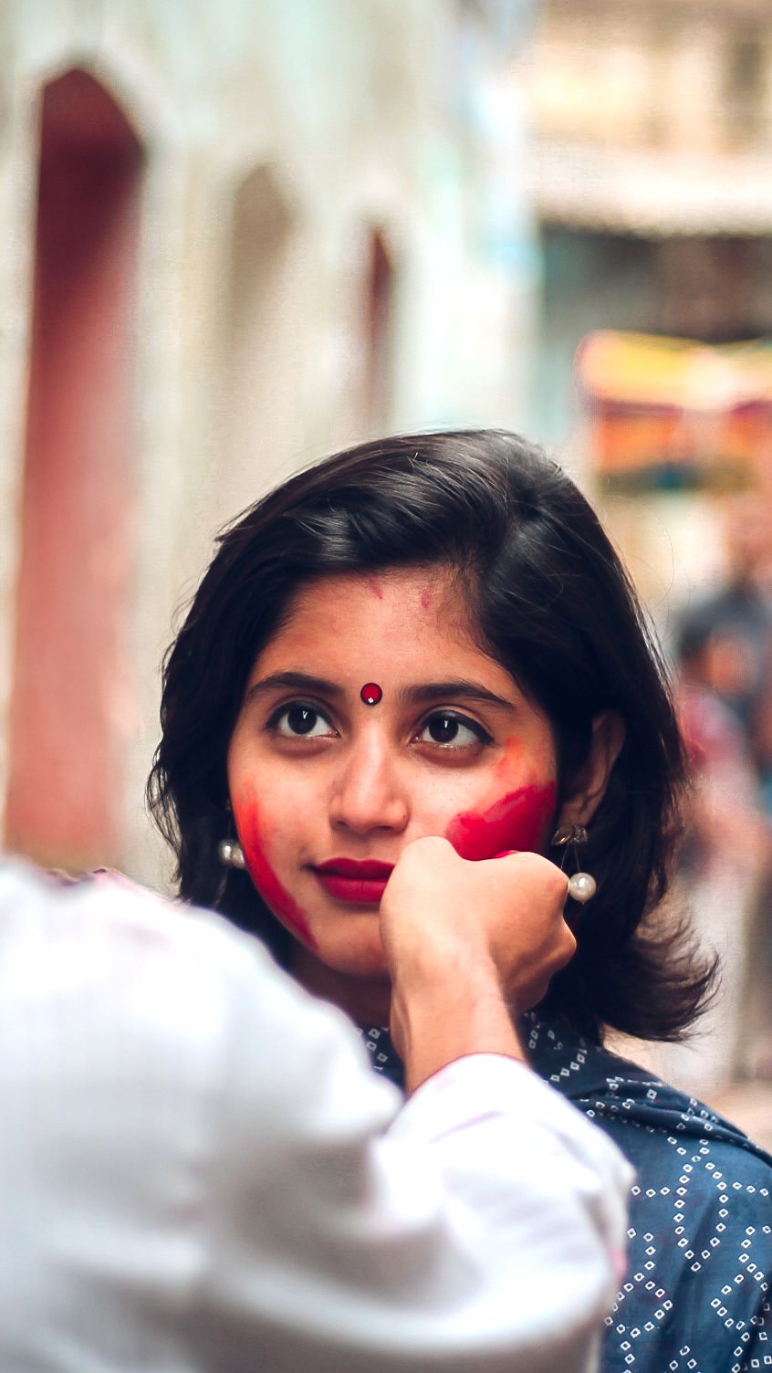 A woman in traditional Indian attire with a red bindi smiles while someone applies color to her cheek during a festive celebration.