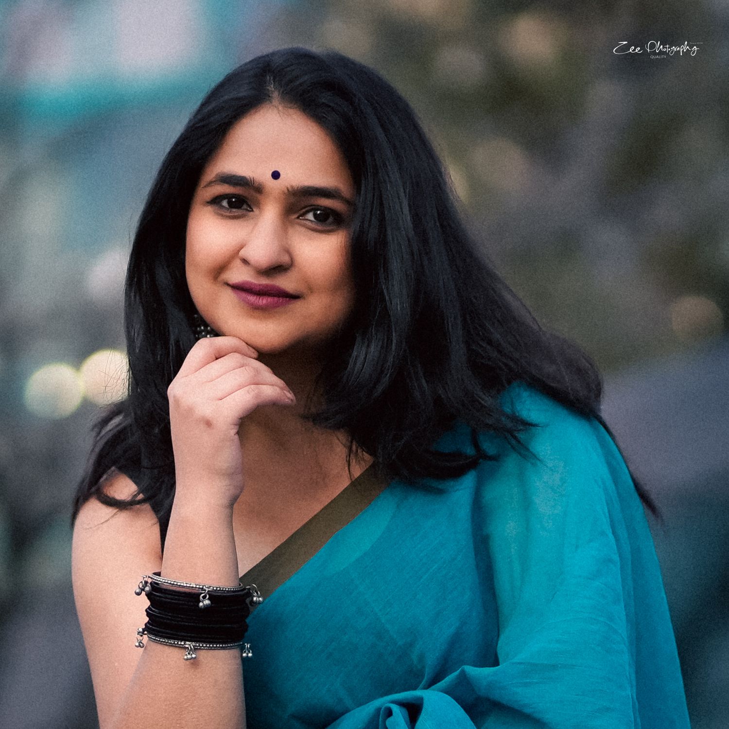A person wearing a turquoise blue saree poses with hand on chin against a blurred background.