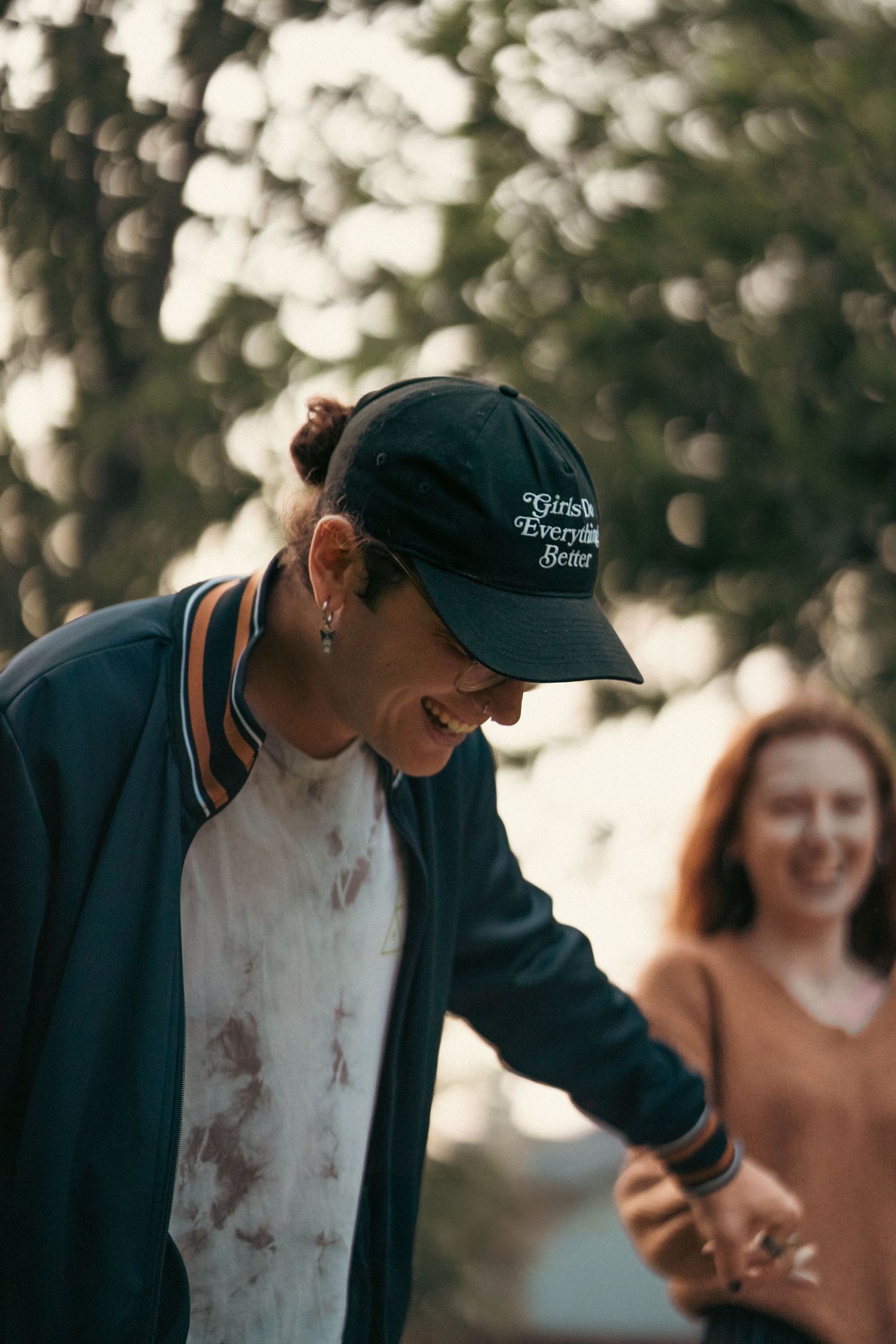 Person wearing a dark green ballcap and jacket laughs with friends outdoors in soft bokeh lighting.