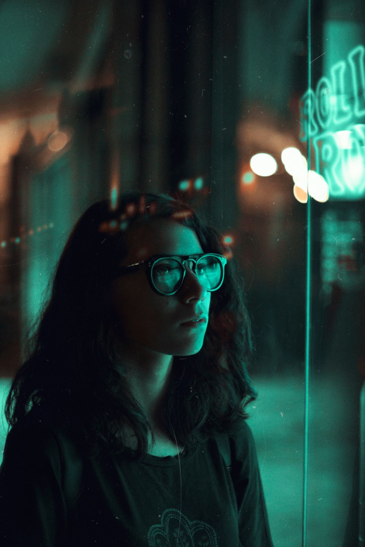 A person wearing glasses illuminated by moody teal neon lighting in an urban nighttime scene.