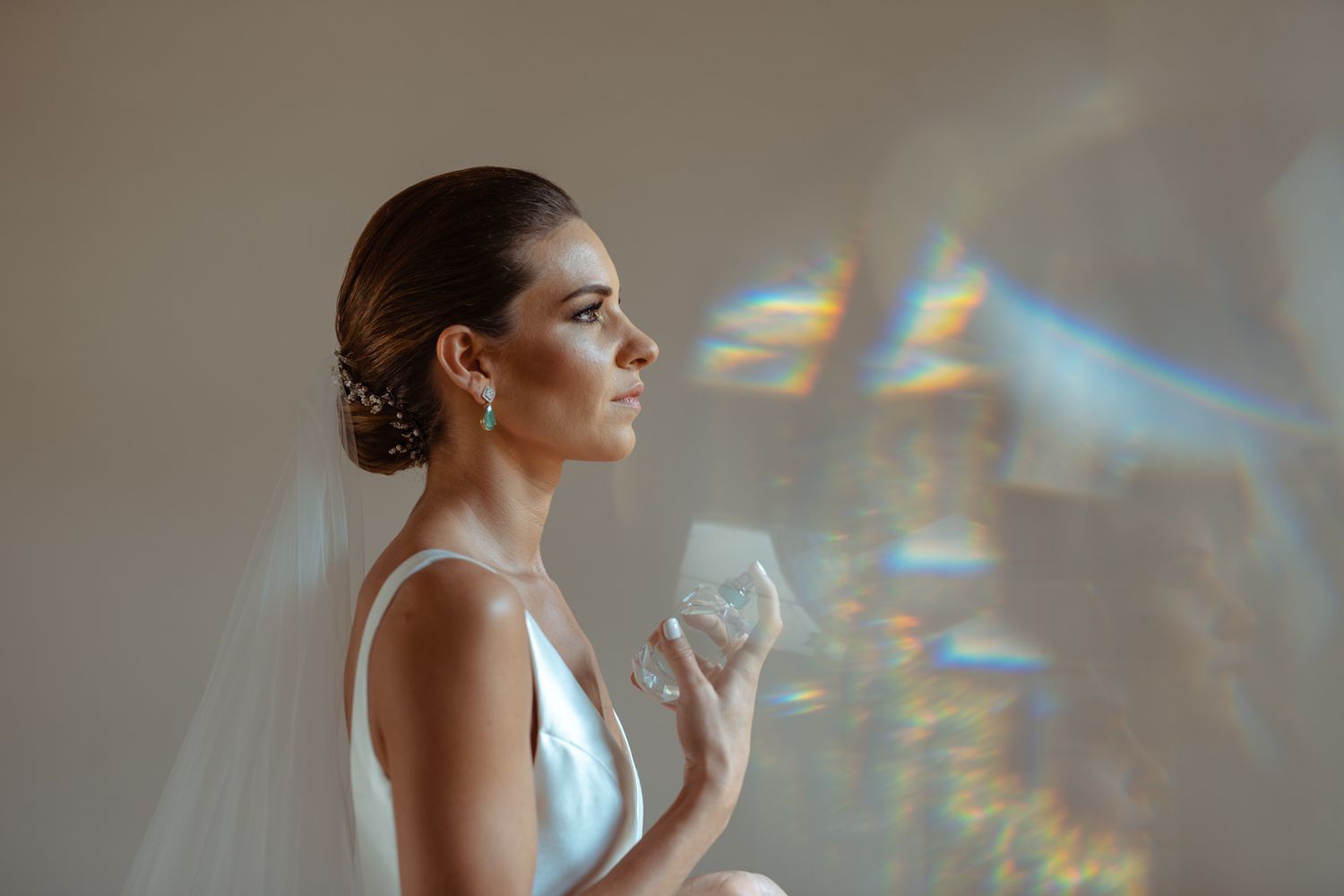 Bride in sleek wedding dress poses with rainbow prism light effects on wall.