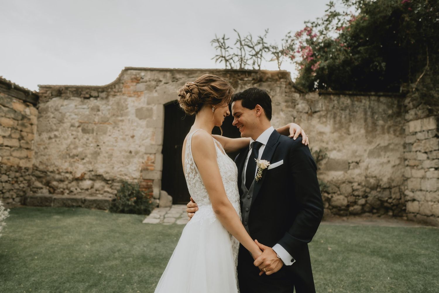 Newlyweds share an intimate moment against an old stone wall in a garden setting.