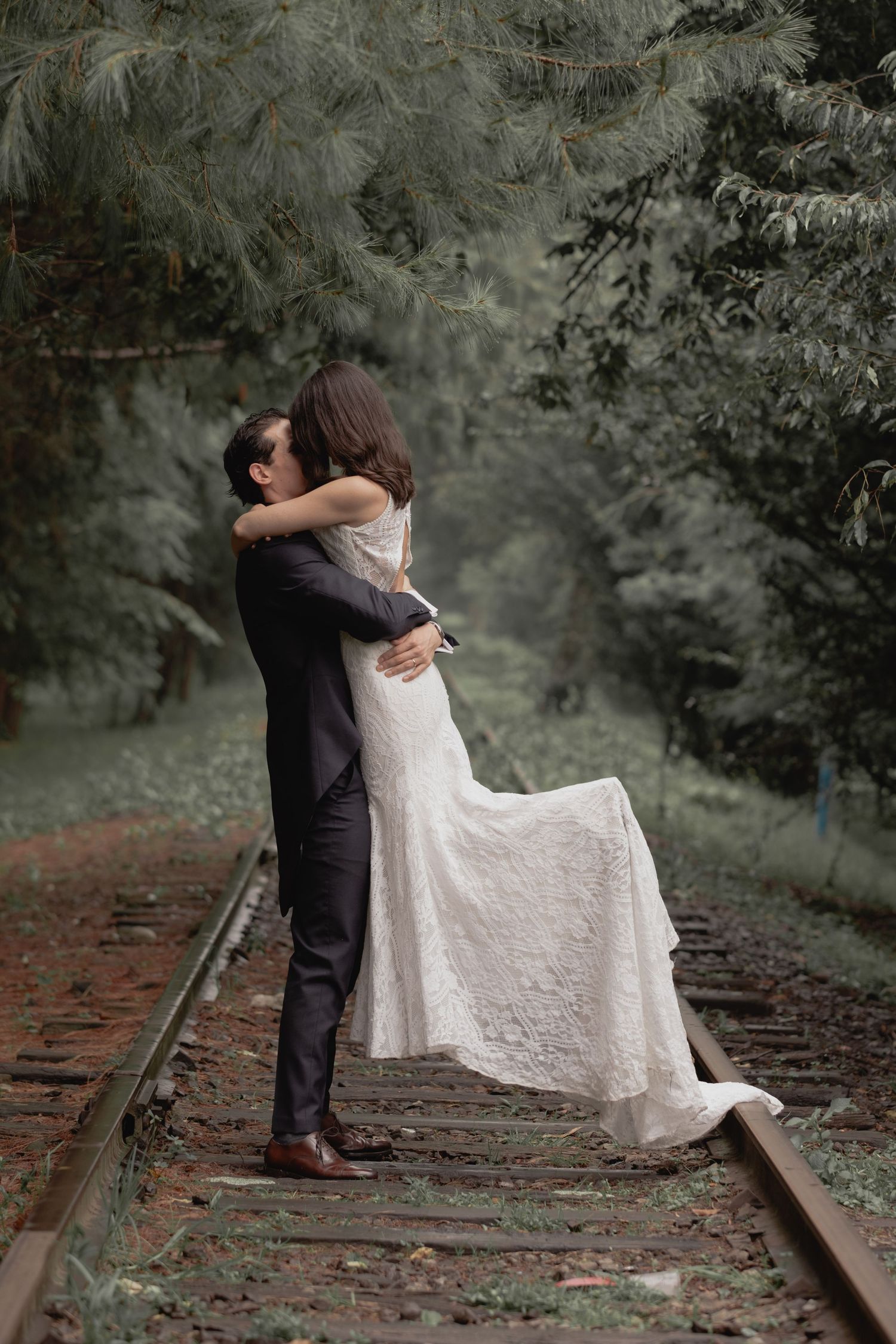 A joyful wedding couple shares an intimate moment on train tracks surrounded by misty trees.