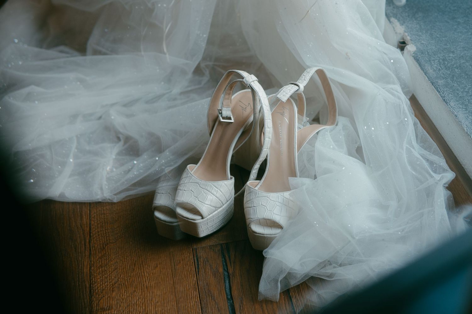 Bridal shoes surrounded by flowing white tulle on dark wooden surface.