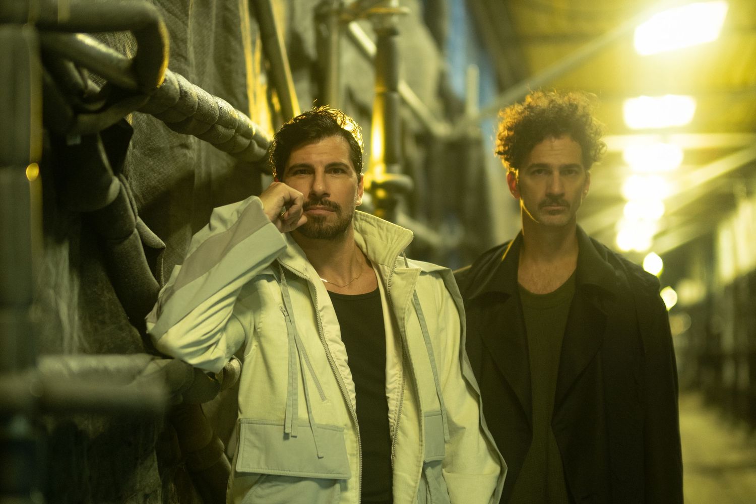 Two people pose in an atmospheric nighttime alley with yellow lighting and urban backdrop.