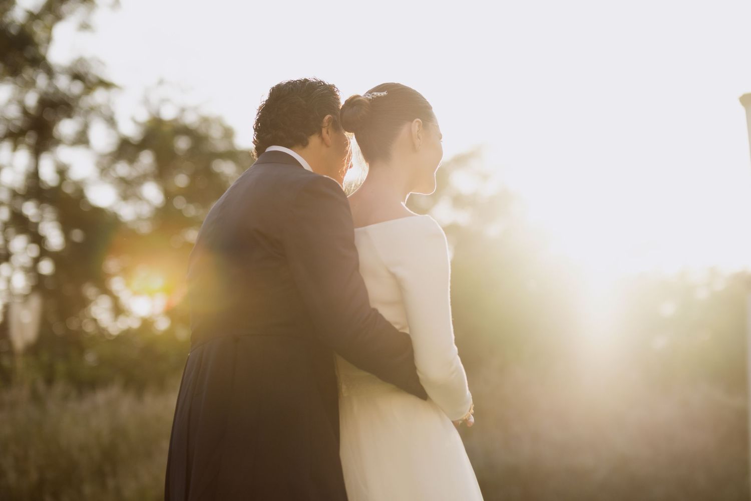 Romantic wedding couple embraces in golden sunset light with dreamy backlit glow.