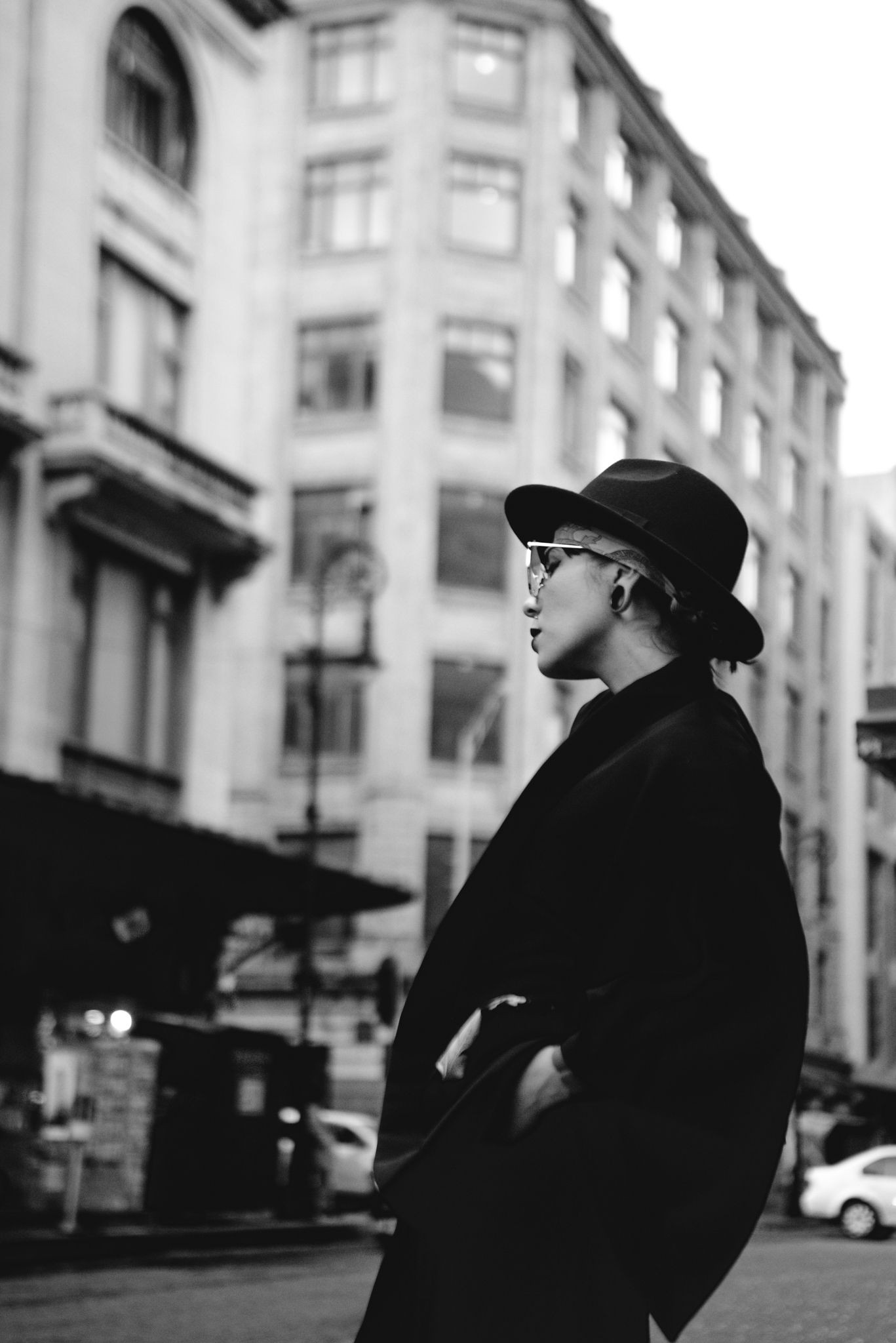 A stylish figure in black coat and hat stands against an urban backdrop of tall buildings.