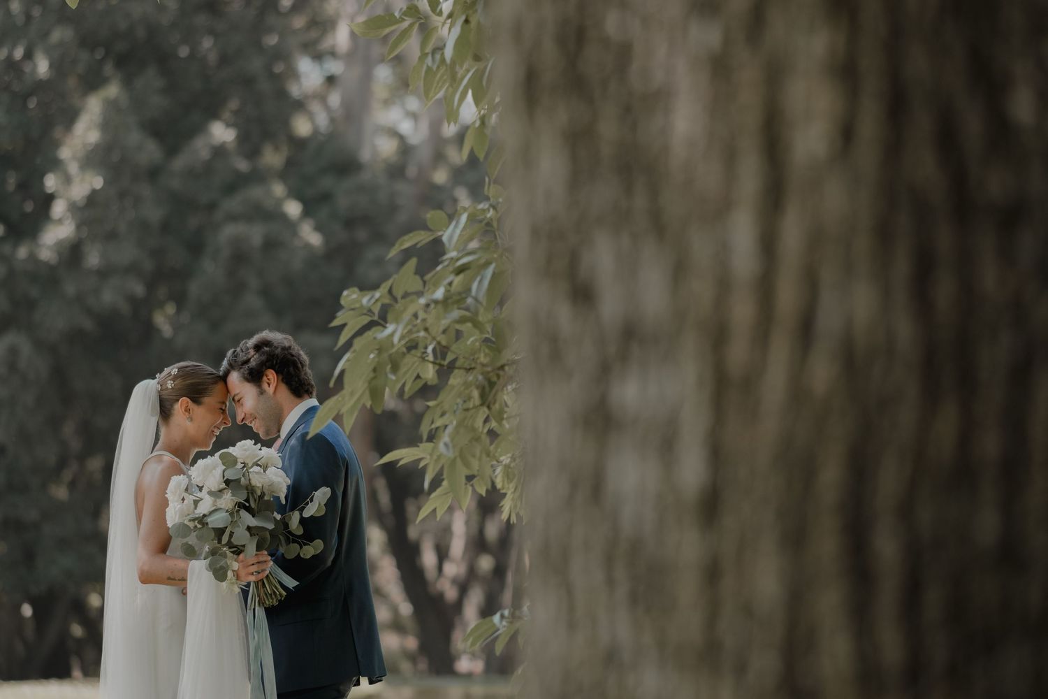 A couple in wedding attire shares an intimate moment outdoors in a lush wooded setting.