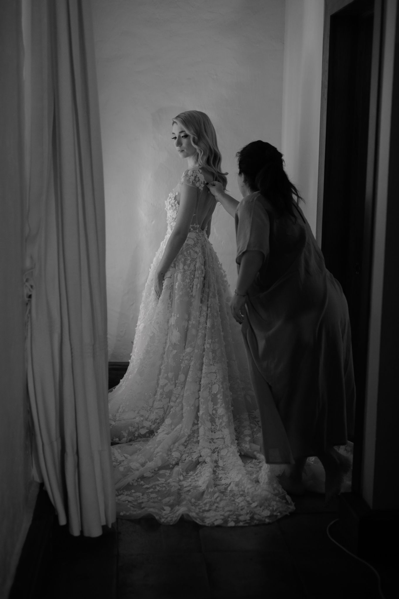 A black and white photograph shows final adjustments being made to an intricate lace wedding gown in a softly lit room.