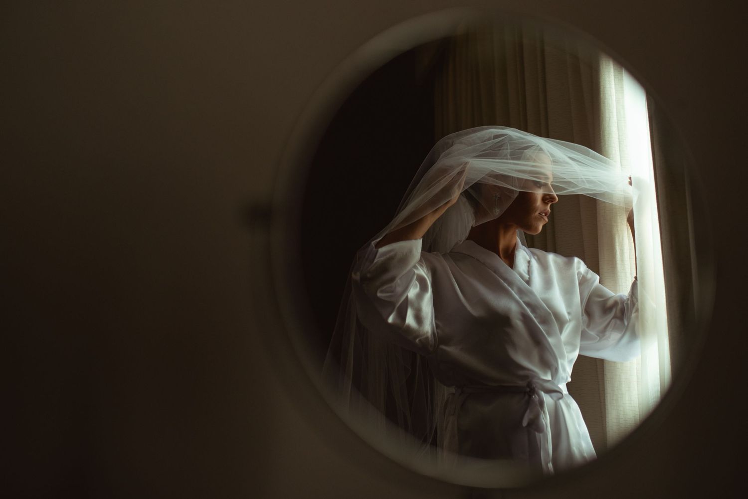 A person in a white robe with flowing material stands by a window in soft, moody lighting viewed through a circular frame.
