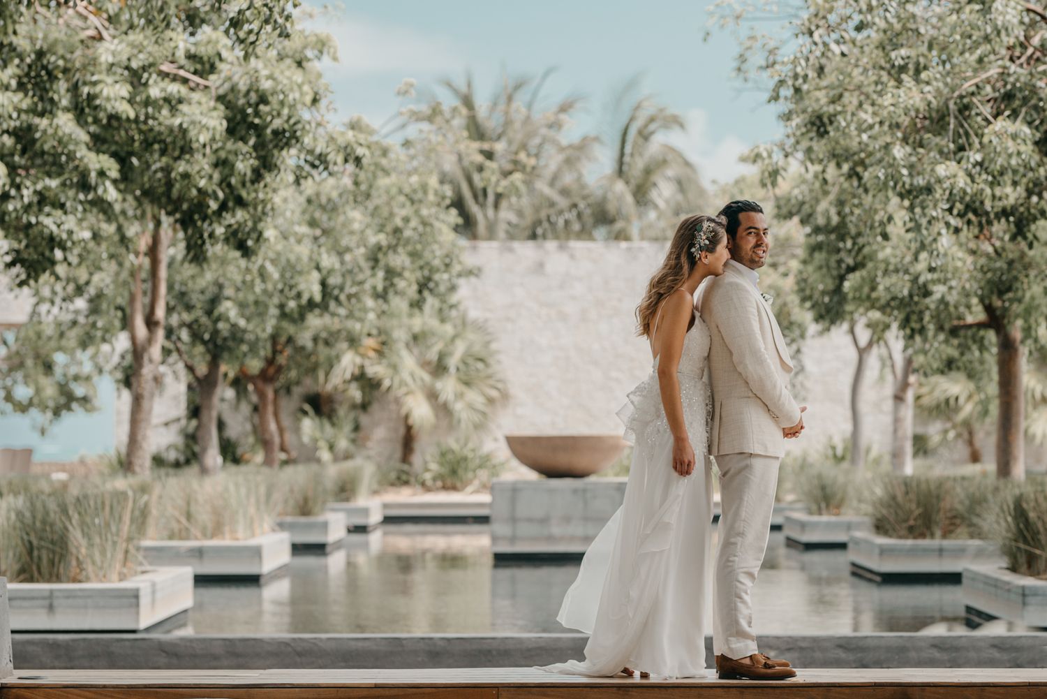 A couple stands back to back in elegant white attire at a luxury resort with palms and reflecting pools.