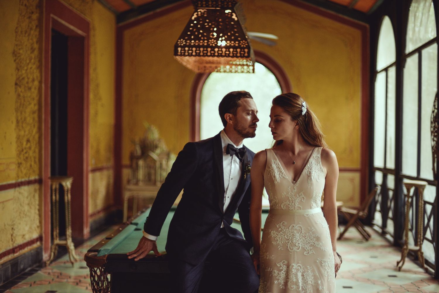 Wedding couple in elegant formal attire share an intimate moment in a vintage indoor setting with arched windows.