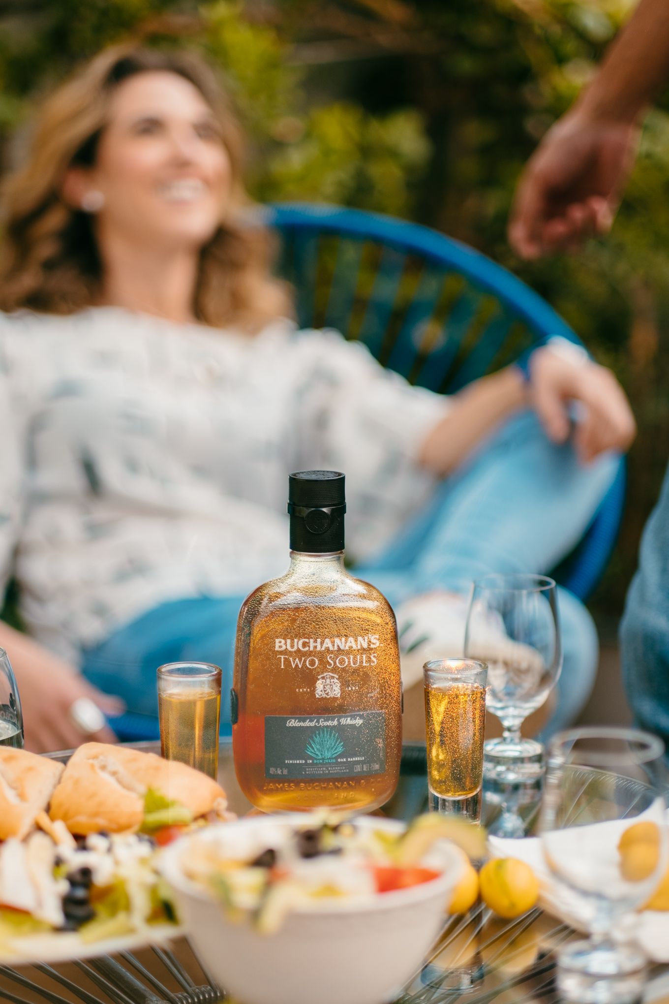 A bottle of Buchanan's whisky sits on an outdoor table with plates of food and drinks during a sunny gathering.