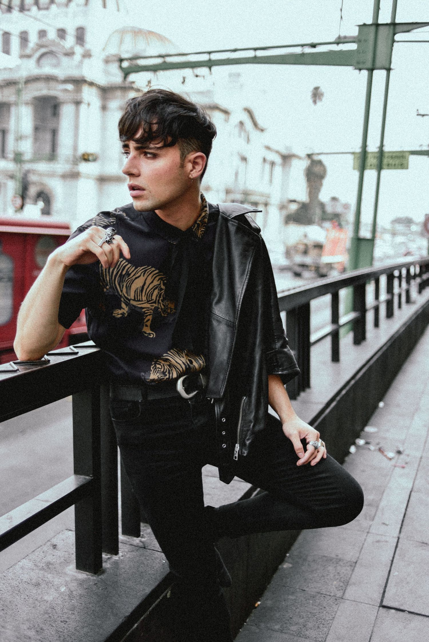 Young man in dark streetwear poses against railing in urban setting.