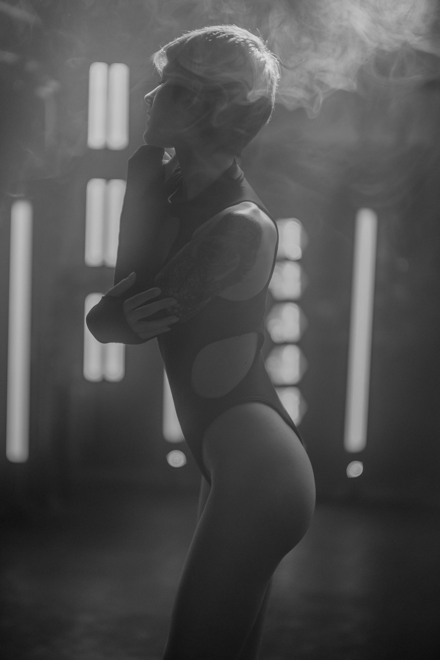 Artistic black and white photograph of a silhouetted figure in a bodysuit posing against bright backlighting in smoky atmosphere.