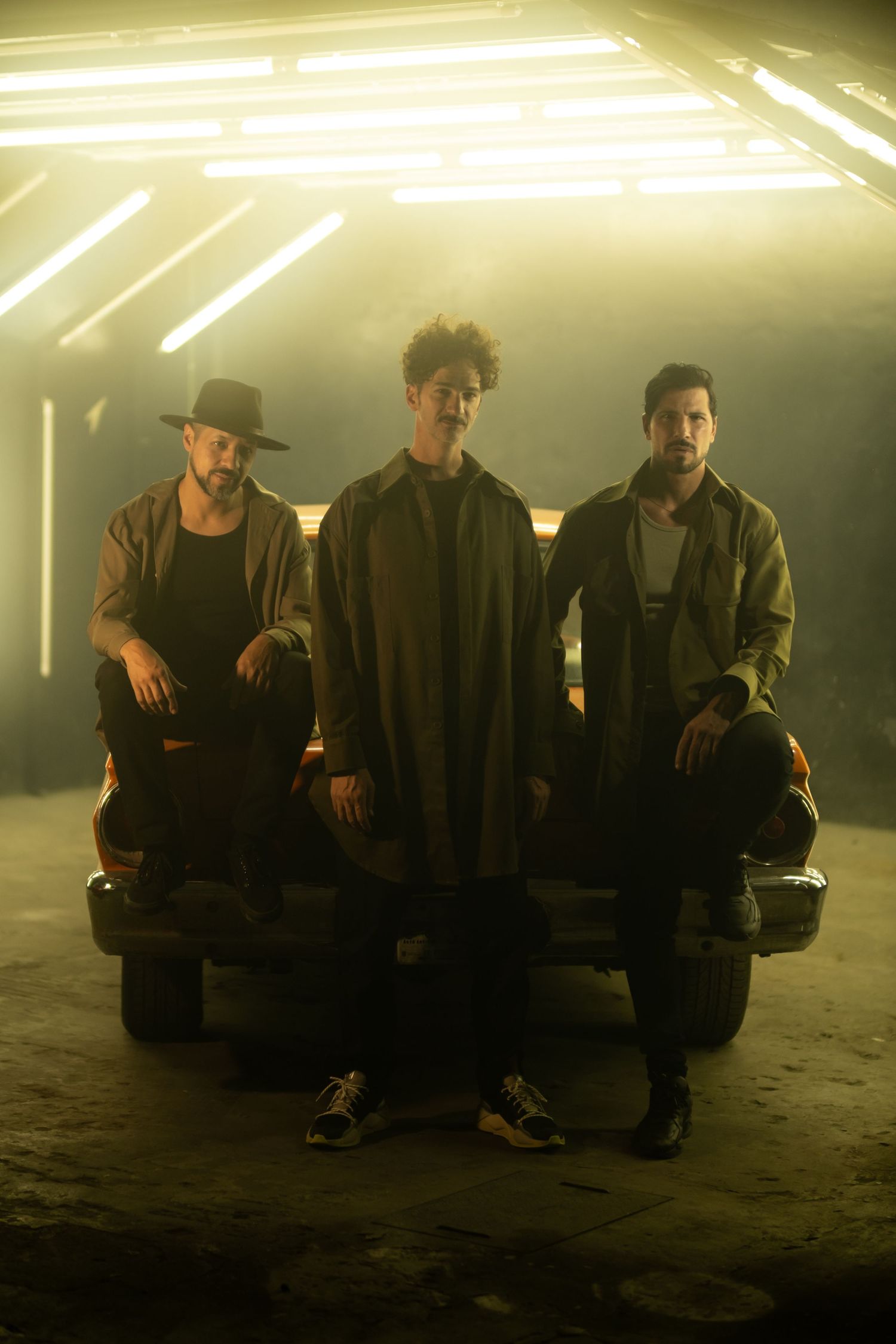 Three musicians pose dramatically under bright overhead lighting on dark stage.