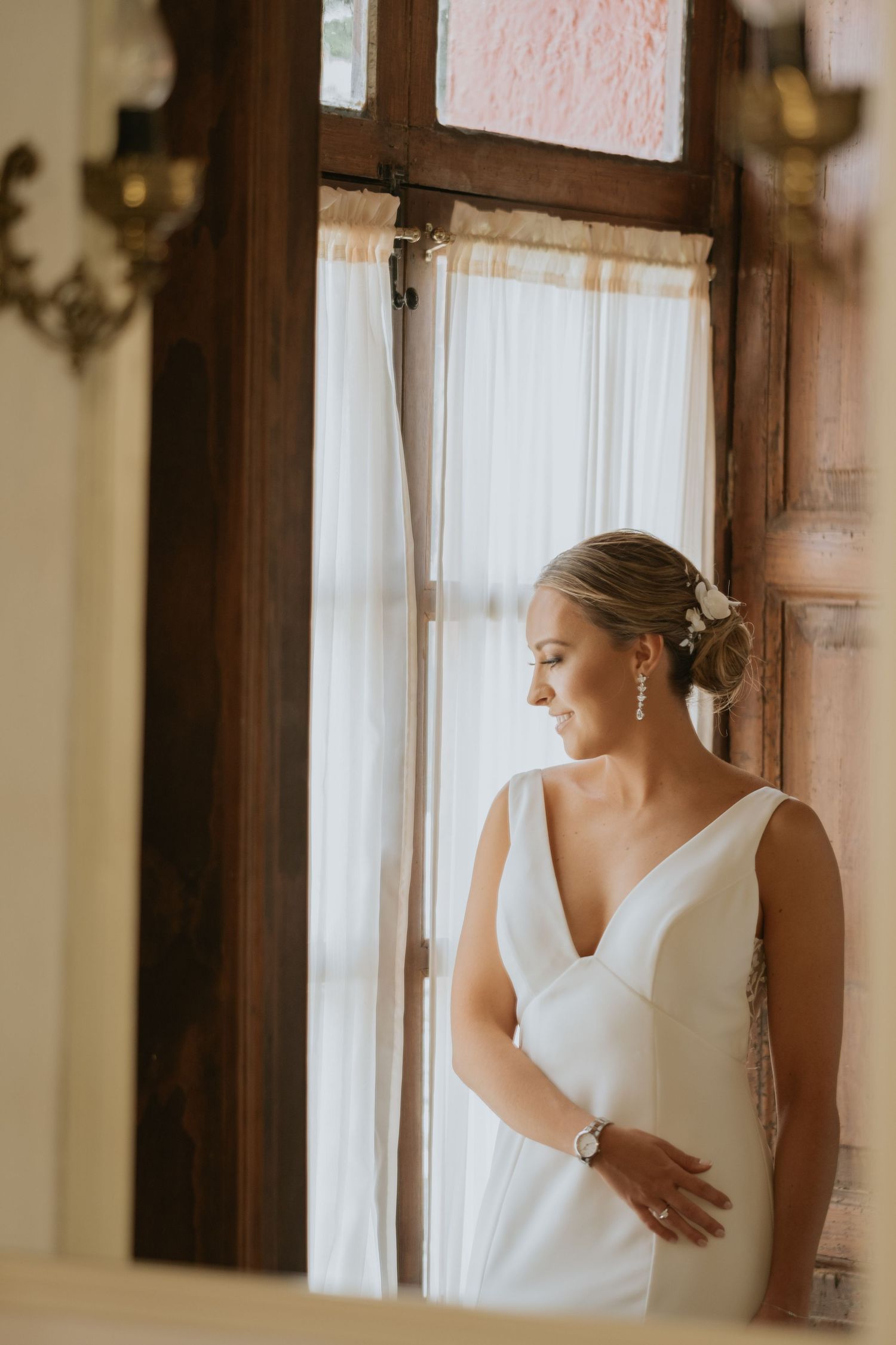 A bride in a classic white wedding dress stands elegantly by a window in natural light.