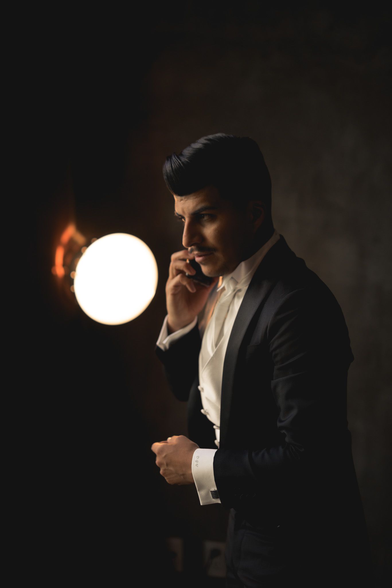 A person in a formal black suit stands in dramatic lighting with a glowing orb light fixture behind them.