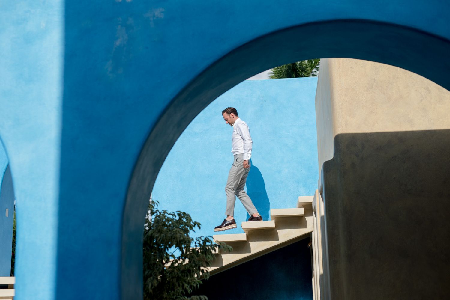 Person walks down white concrete stairs framed by a vibrant blue archway against a sunny Mediterranean backdrop.
