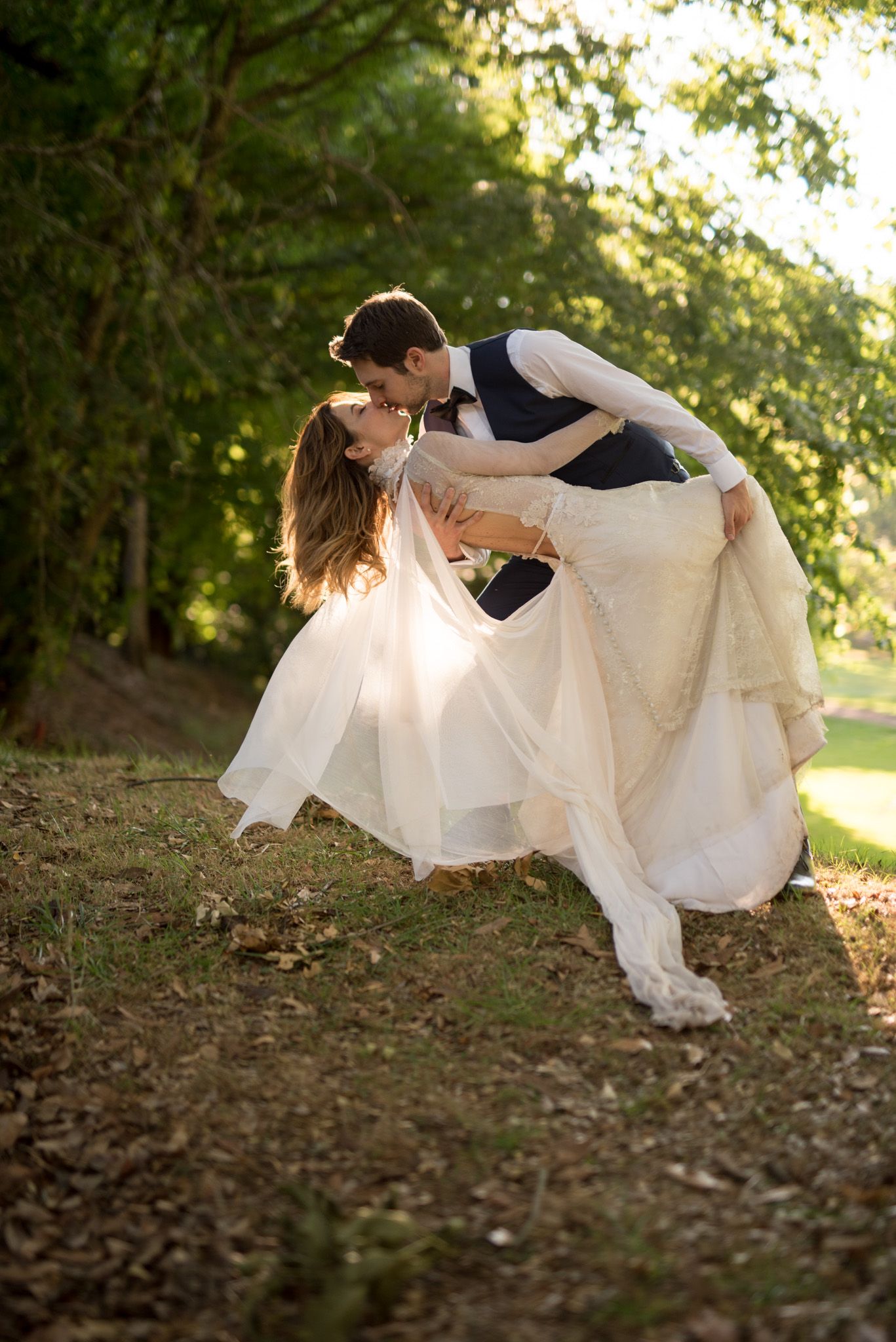 A romantic outdoor wedding photo showing a graceful dip pose against a sunlit natural woodland backdrop.