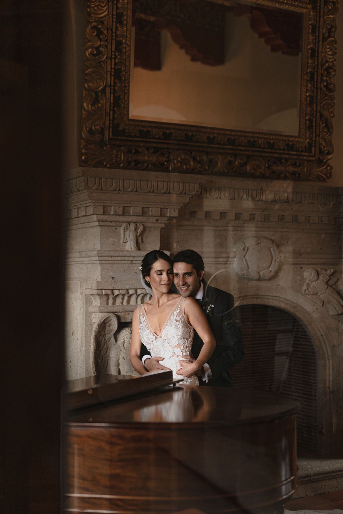 A romantic couple in formal attire poses together near an ornate fireplace and mirror in a dimly lit vintage room.