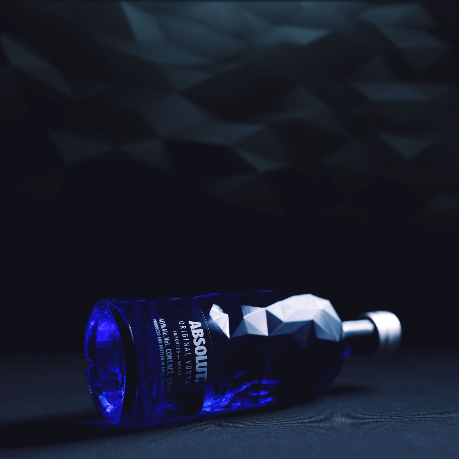 Artistic still life photo of a blue illuminated glass bottle lying sideways on dark reflective surface.