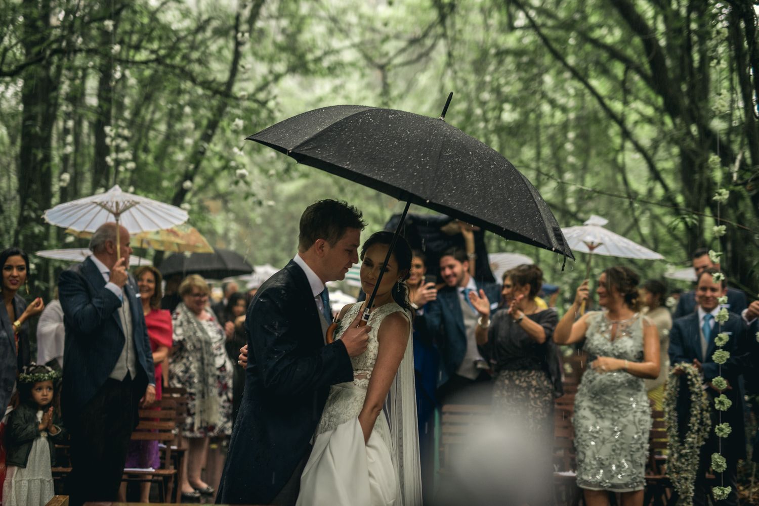 A wedding ceremony in a lush forest setting during light rain with guests holding umbrellas.