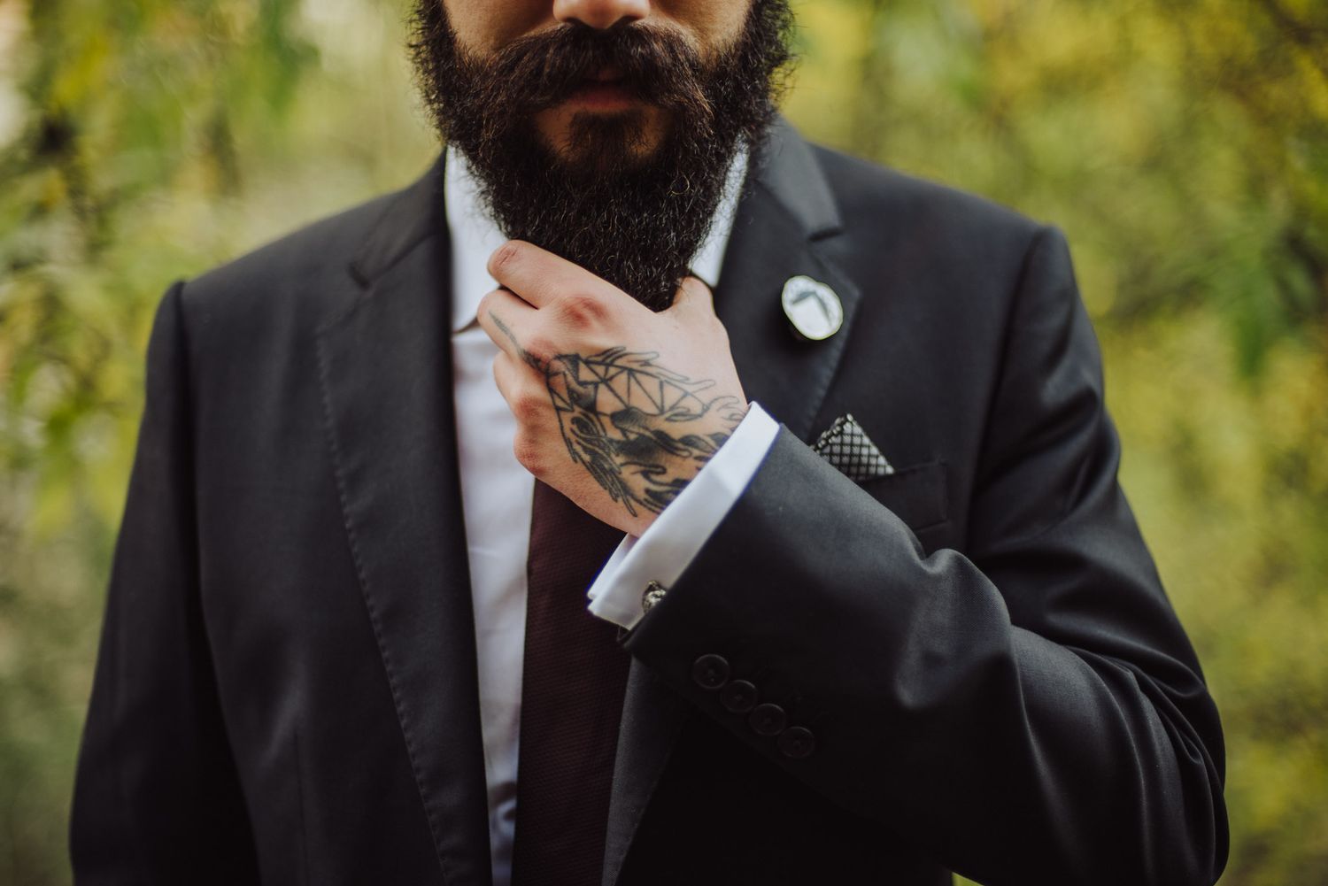 Close-up of groom in black suit adjusting sleeve cuff showing detailed tattoo artwork and groomed beard.