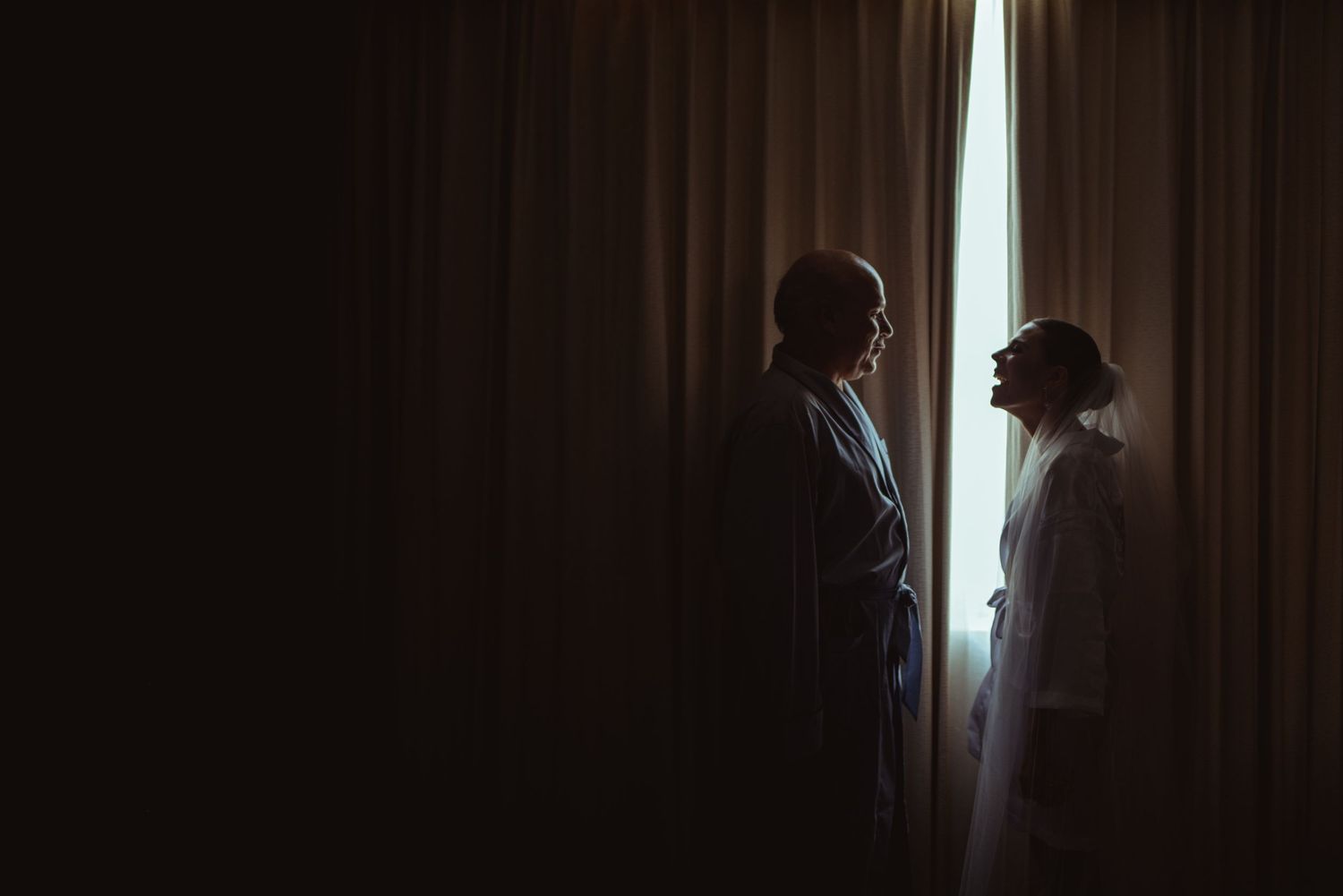 Two silhouetted figures stand in dramatic lighting near curtained window creating a moody atmospheric scene.