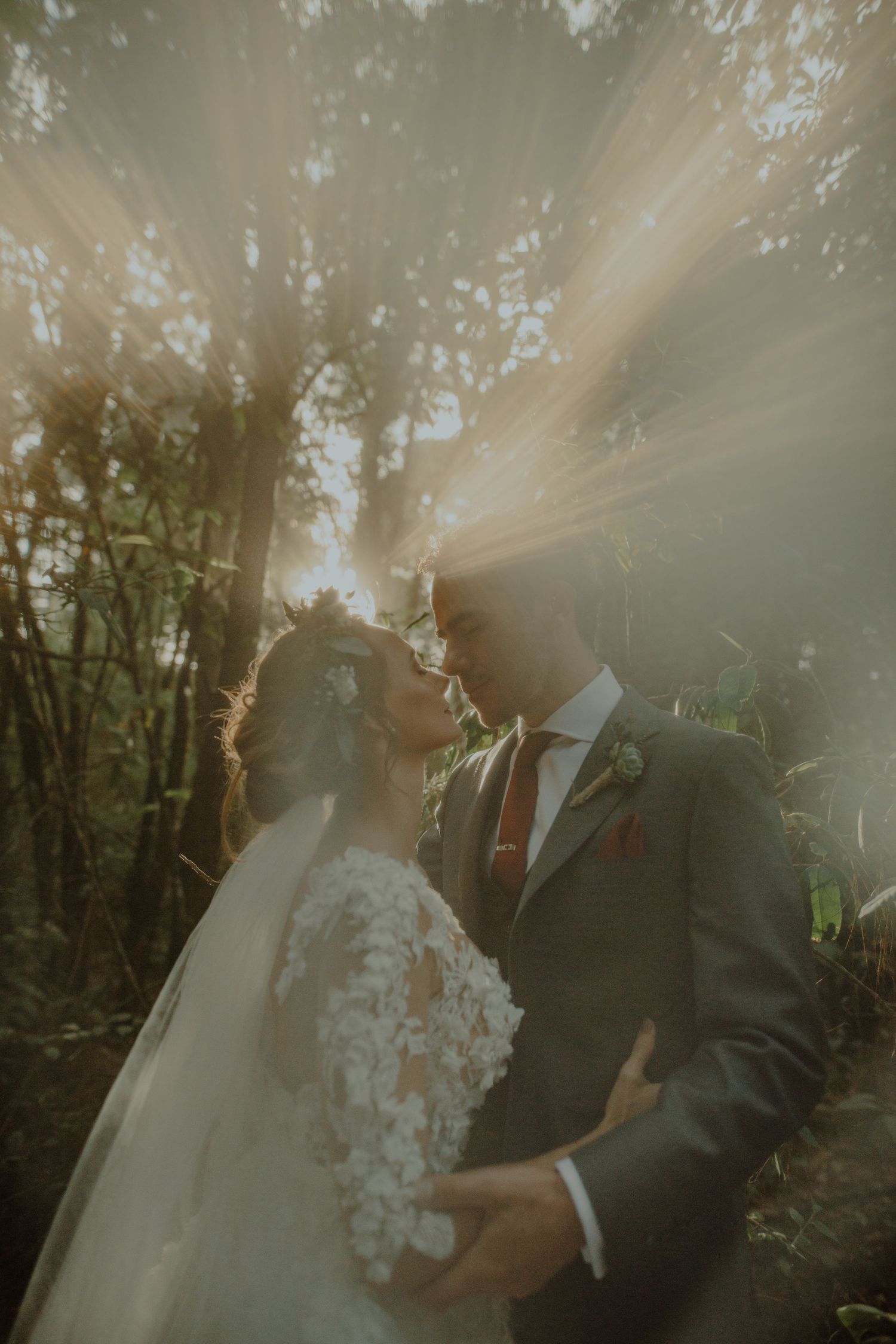 Ethereal sunbeams shine through forest trees as newlyweds share an intimate moment in lace dress and dark suit.
