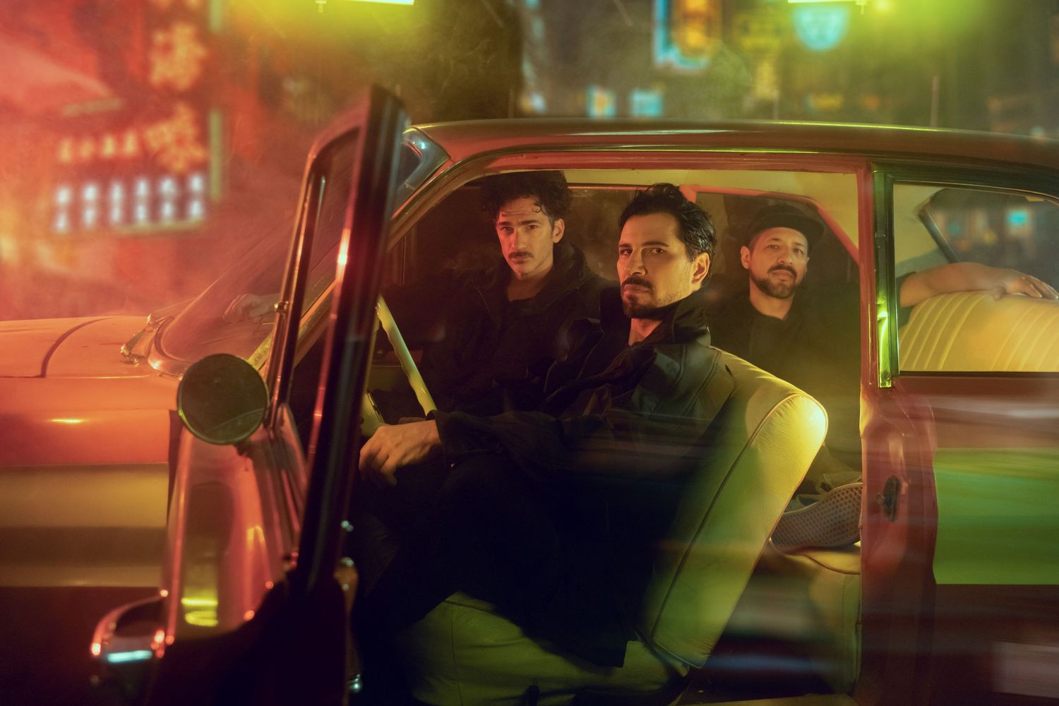 Three occupants sit inside a car at night with neon city lights glowing in the background.