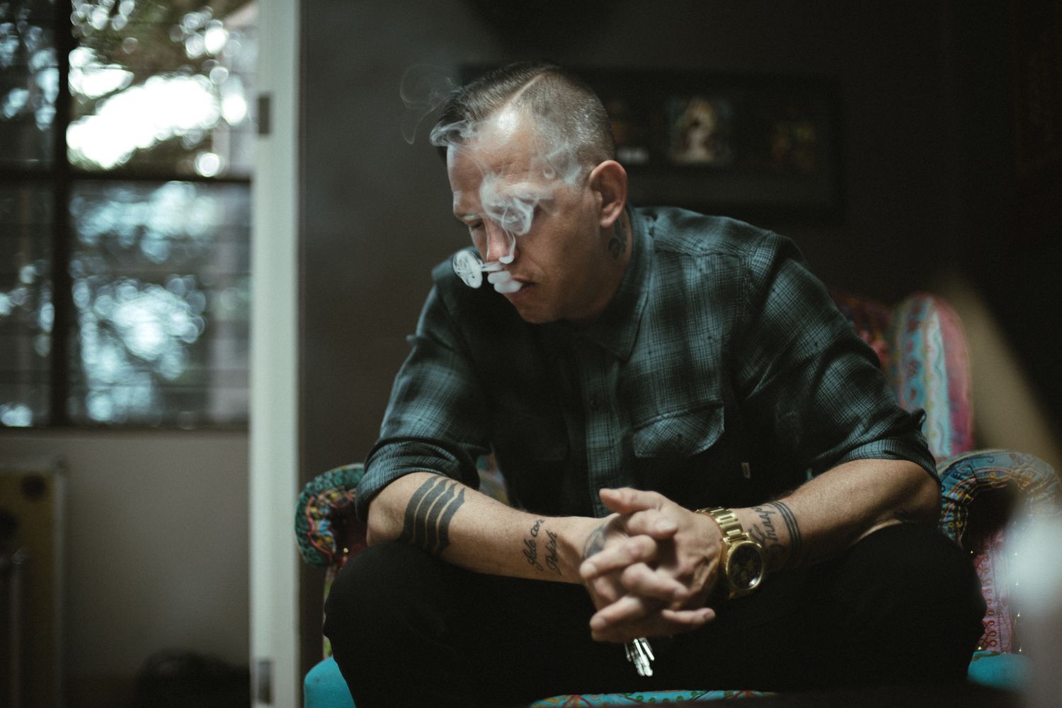 A contemplative figure in glasses and plaid shirt sits in a shadowy indoor setting.