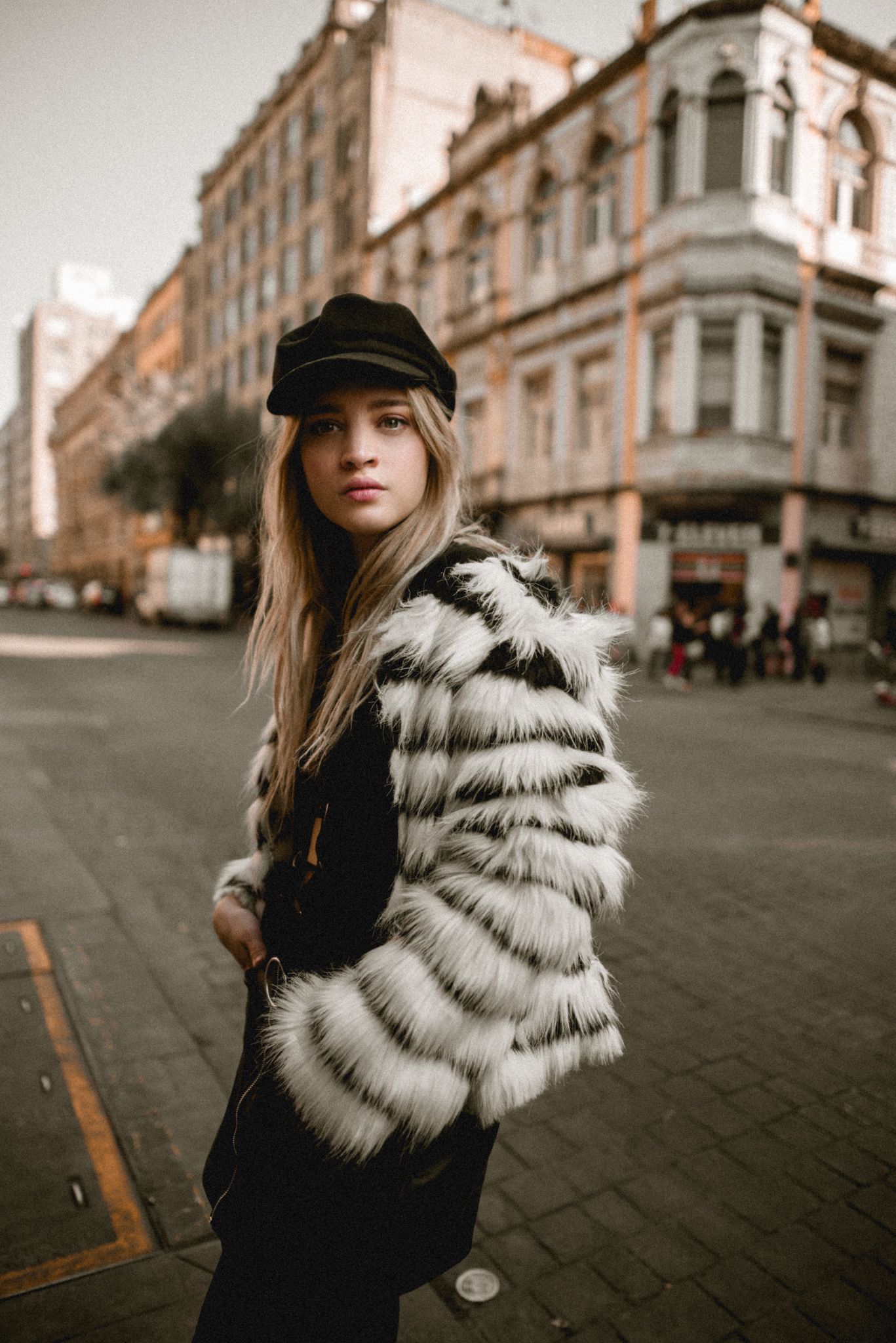 Street fashion model wearing striped fur coat and black cap poses against urban architecture background.