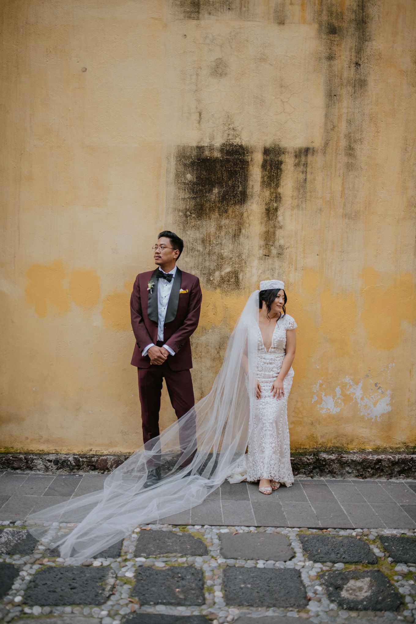 Wedding couple poses against weathered yellow wall with long flowing veil.