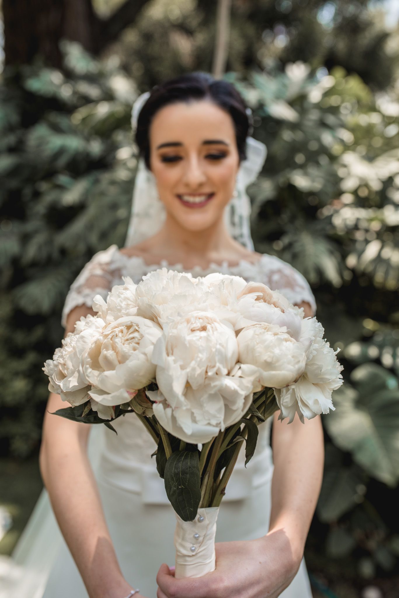 An elegant white peony bridal bouquet with satin ribbon wrap against a garden backdrop.