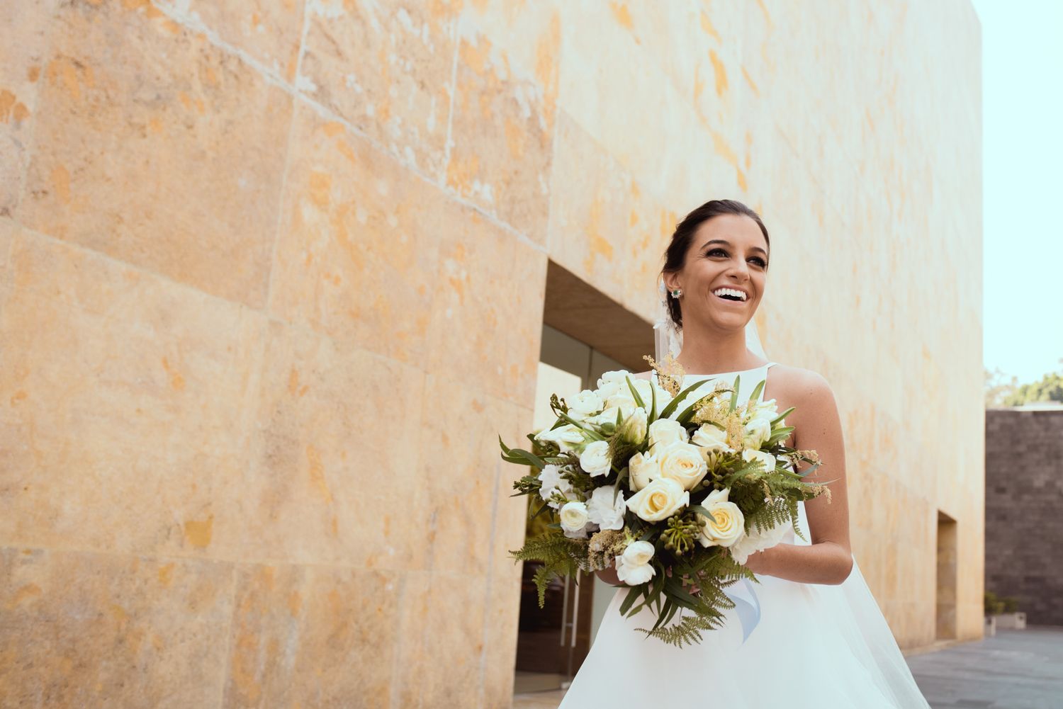 A person in a white wedding dress holds a bouquet of white flowers while standing against a beige stone wall.