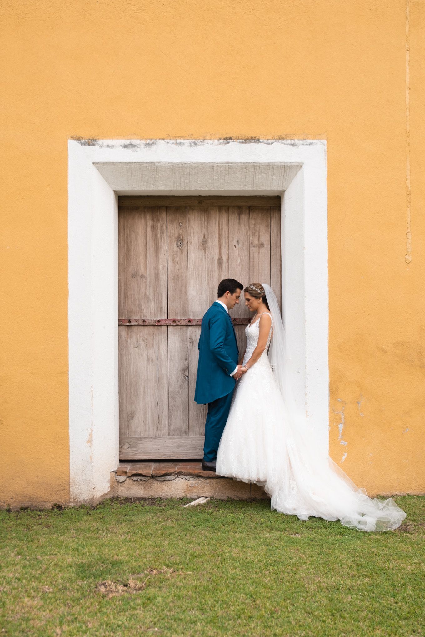 A romantic wedding portrait against a rustic wooden door and yellow wall showing a couple in formal attire.