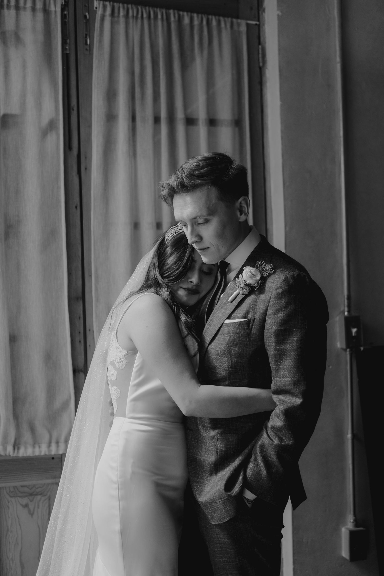 Intimate black and white portrait of newlyweds sharing a tender embrace by a window with sheer curtains.