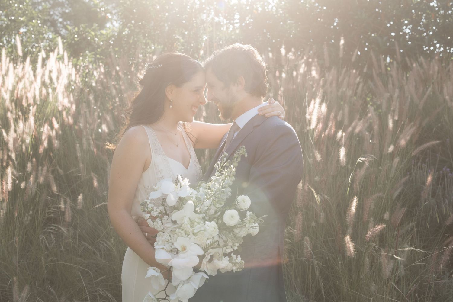 Newlyweds share intimate moment surrounded by sunlit ornamental grasses.