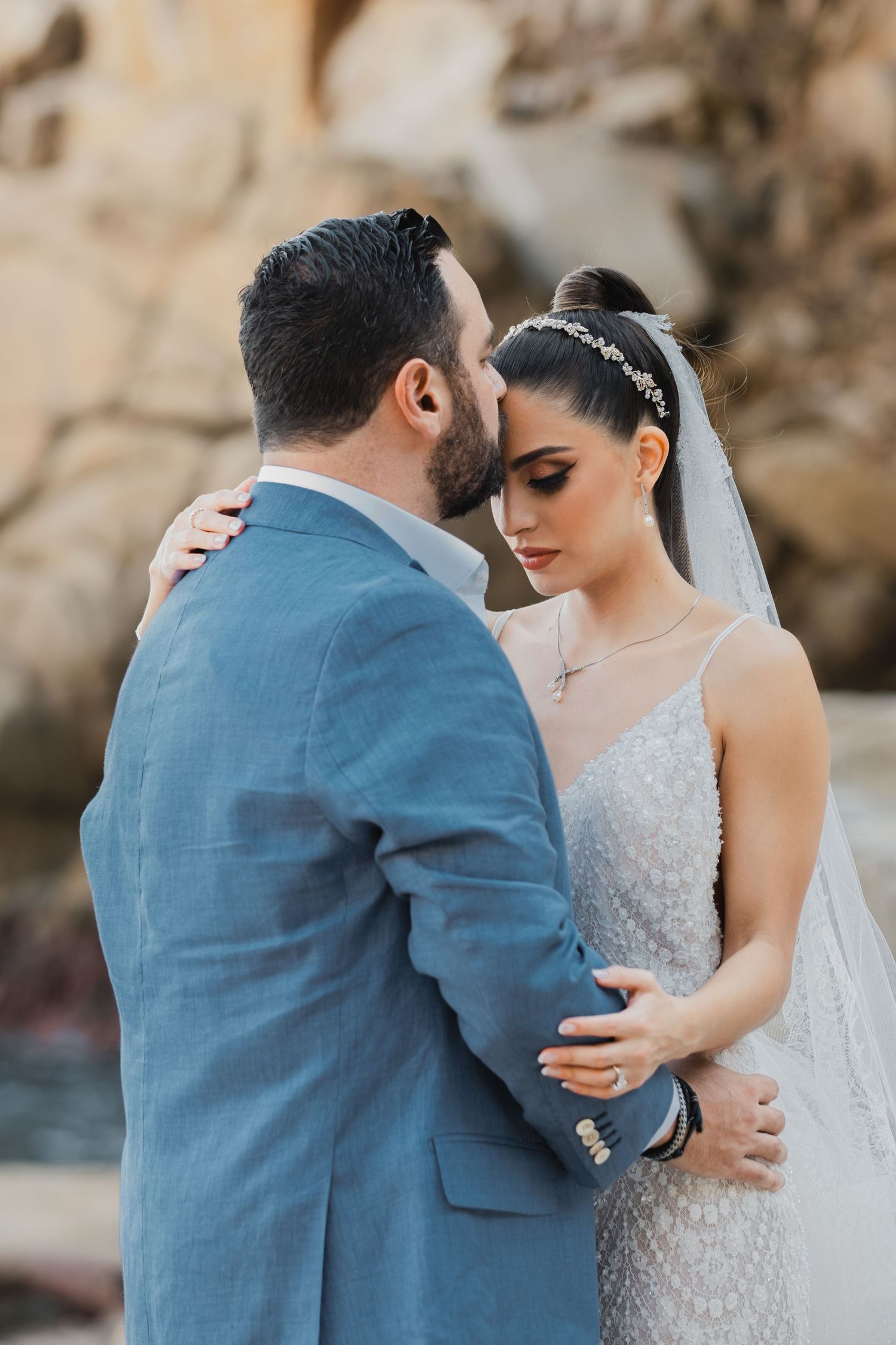 An intimate moment between newlyweds with a lace wedding dress and blue suit against a rocky backdrop.